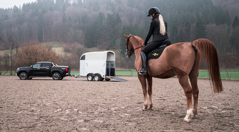 Rückansicht: Reiterin auf Pferd sitzend, im Hintergrund befindet sich ein Gespann aus einem PKW mit angehängtem Pferdeanhänger | © Humbaur GmbH